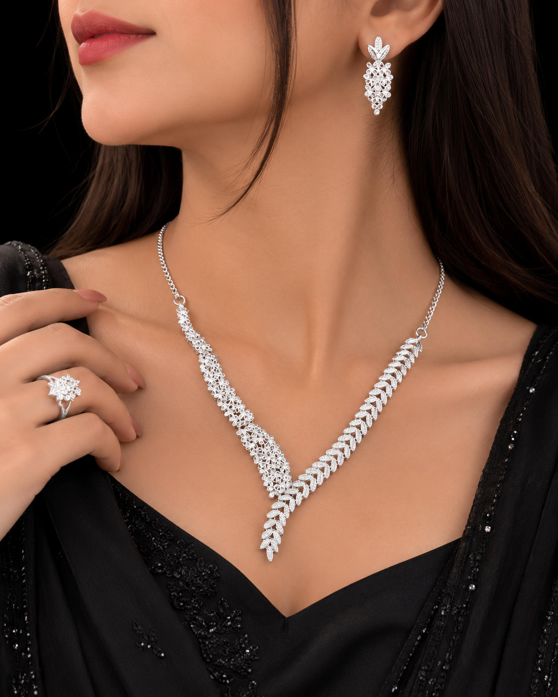 A close-up of a woman wearing a sparkling silver-tone necklace and matching drop earrings, with a matching ring on her finger and a black embellished dress against a dark background.